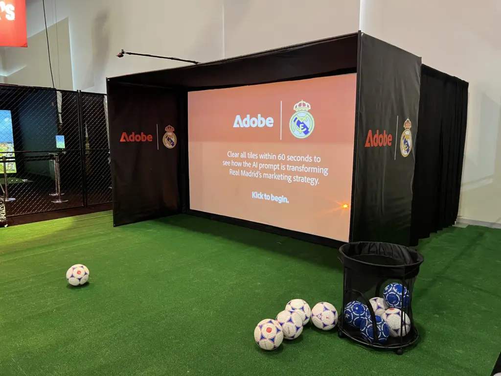 Adobe and Real Madrid branded soccer simulator enclosure with the Kick to begin start screen and balls on the turf at Adobe Summit 2026, Las Vegas