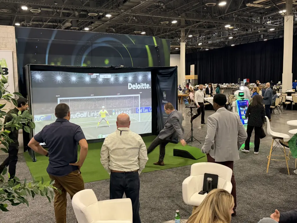Deloitte guests watching a player kick at the branded soccer simulator, with the AI photo booth robot in the background, Venetian Expo, Las Vegas
