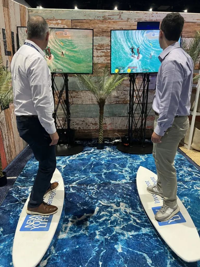 Two players racing on the surf simulators with scores on screen at the New York Life booth, San Diego Convention Center