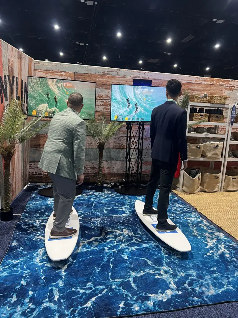 Two attendees in suits riding New York Life branded surf simulators in a beach-themed booth at LPL Focus 2026, San Diego Convention Center