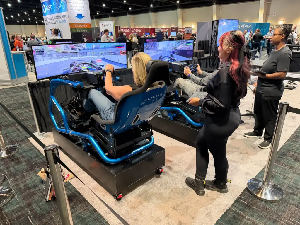 Two attendees racing head-to-head on F1-style racing simulators while a Los Virtuality host coaches them at NISC Member Information Conference 2026, MGM Grand Marquee Ballroom