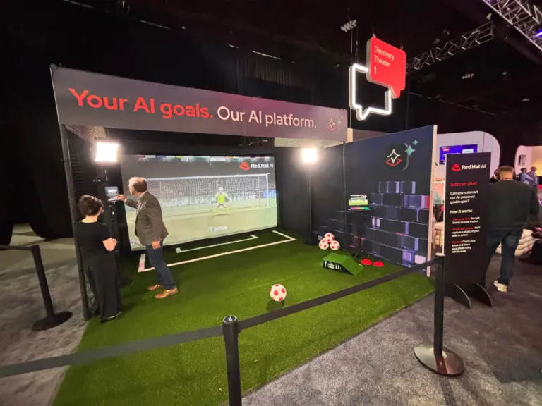 Red Hat AI branded soccer simulator booth with a Your AI goals banner, turf pitch and an attendee taking a shot at Red Hat Summit 2026, Georgia World Congress Center, Atlanta